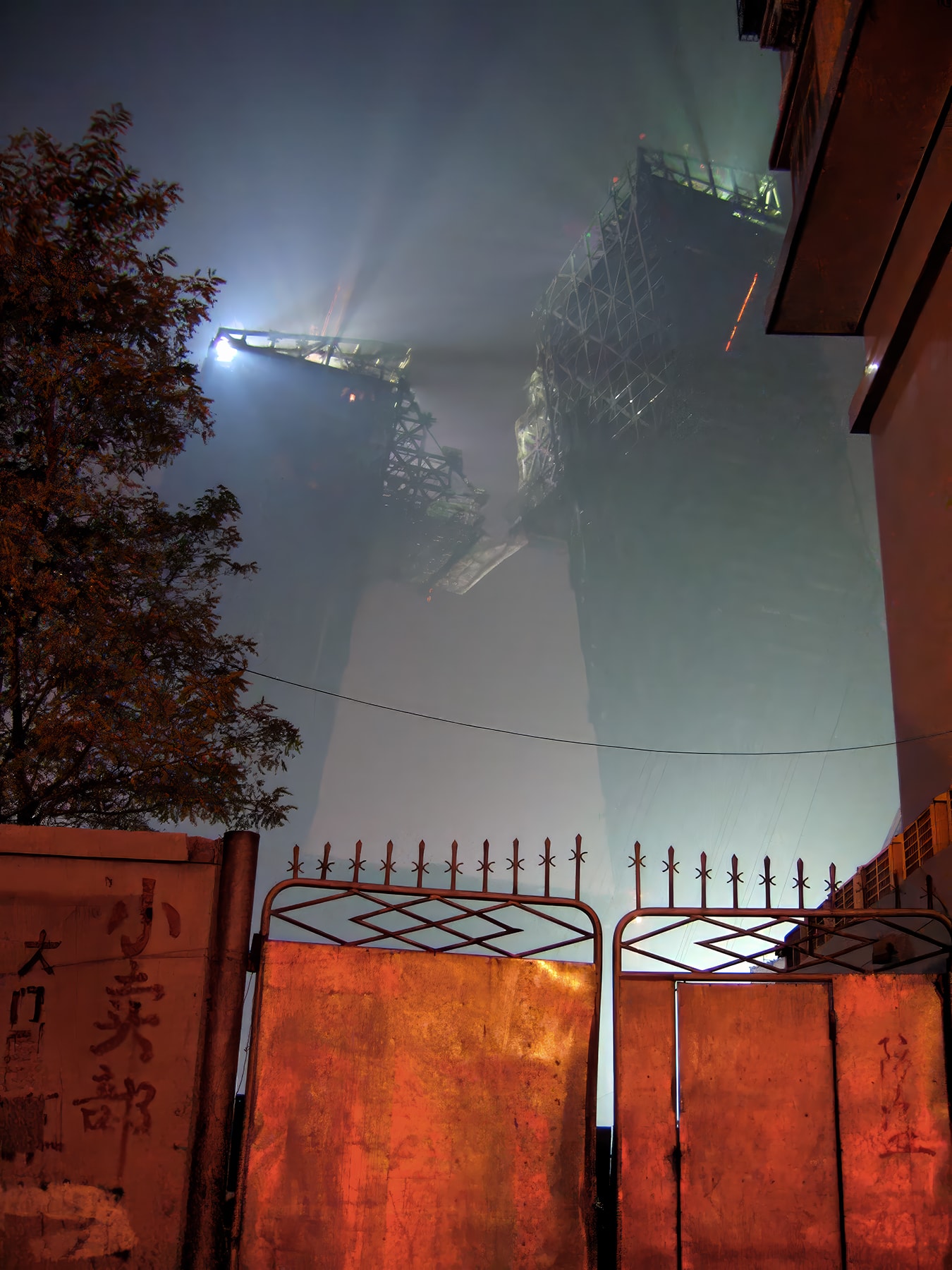 Construction cranes in fog above rust-red iron gates with Chinese signage at night