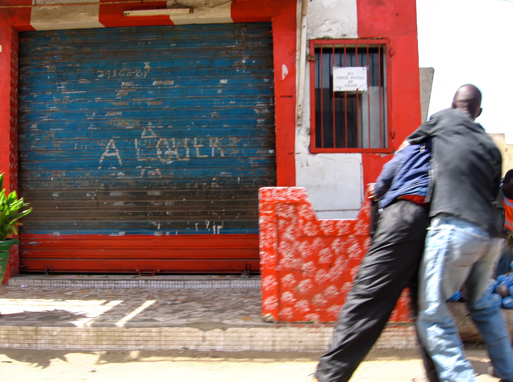 Men walking past red storefront with blue shutter, Dakar